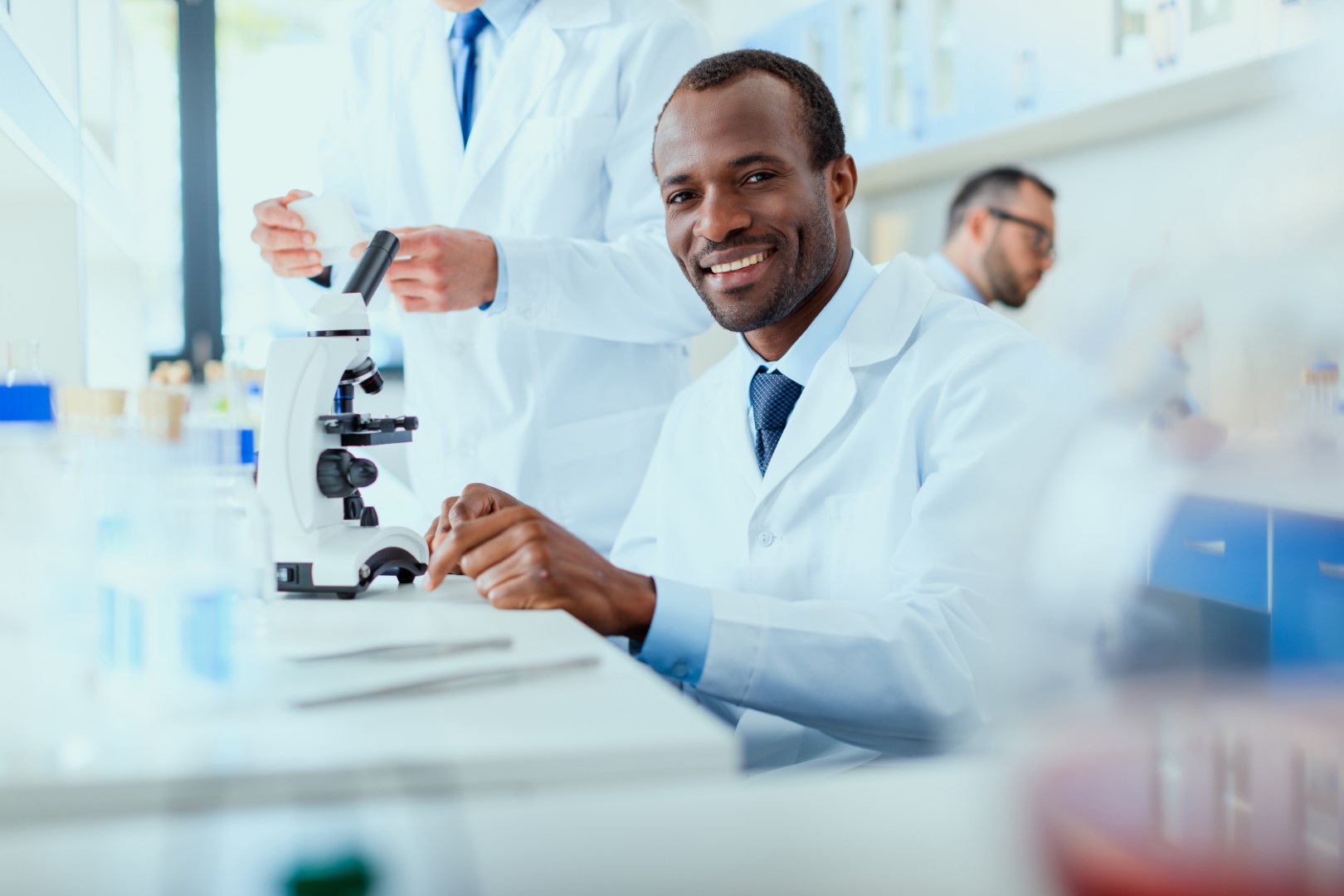 A group of young men working in a laboratory. The man in the center of the image is sitting in front of a microscope and smiling at the camera.