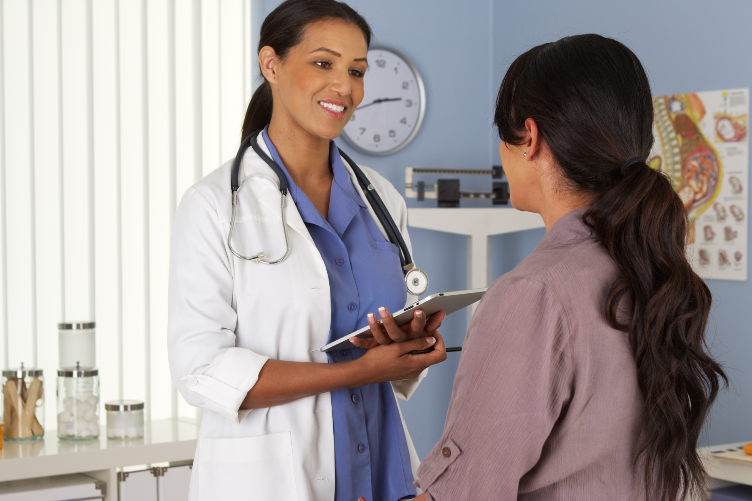 A female gynecologist holding a tablet talking to a woman in an exam room.